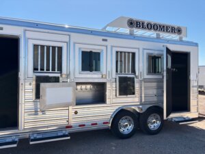 A shiny silver Custom Show Trailer with three barred windows, a side compartment, and two open doors. The trailer has dual wheels, a BLOOMER sign on top, and is parked outdoors under a clear sky. Perfect for horse and livestock trailer sales. -Stillwater Trailer Sales