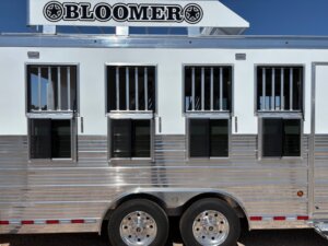 A silver and white horse trailer with four barred windows, two axles with chrome wheels, and a Bloomer sign on top, parked outdoors under a clear blue sky—perfect for those seeking new and used trailers or expert horse and livestock trailer sales. -Stillwater Trailer Sales