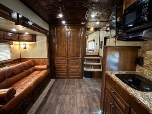 Interior of an RV featuring a brown leather sofa, wood paneled cabinets and walls, a small kitchen with sink and microwave, and steps to a raised section—showcasing Living Quarters custom design found in top horse and livestock trailer sales. -Stillwater Trailer Sales