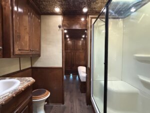 A bathroom with wood-paneled walls, a granite countertop sink, a toilet, and a glass-enclosed shower—features often found in custom show trailers. Beyond the shower, a hallway leads to cabinetry and a bed, all brightly lit by ceiling lights. -Stillwater Trailer Sales