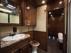 A bathroom with a marble countertop sink, a toilet with a dark seat, wooden cabinets, and beige walls—showcasing the luxury options available in our Living Quarters custom horse and livestock trailer sales. The room leads to a cozy bed area with wood paneling and recessed lighting. -Stillwater Trailer Sales