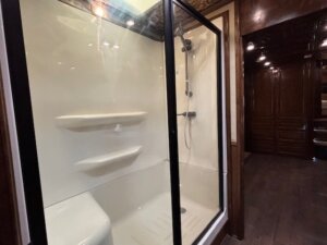 A glass-enclosed shower with two built-in corner shelves and a handheld showerhead, adjacent to a hallway with dark wood paneling and cabinetry—perfect for custom show trailer interiors. -Stillwater Trailer Sales