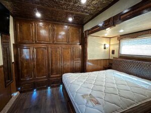 A bedroom with a large wooden wardrobe, wood-paneled walls, a patterned headboard, and an unmade mattress. Ceiling lights glow as natural light enters through a window. A TV is mounted in the corner—ideal for relaxing after browsing custom show trailers online. -Stillwater Trailer Sales