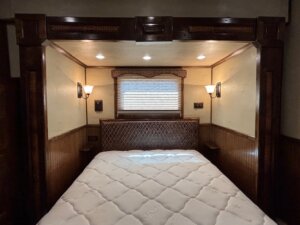 A neatly made bed with a white mattress sits in a cozy, wood-paneled alcove. Overhead recessed lights and wall-mounted reading lamps create a warm retreat—perfect for Living Quarters custom options in horse and livestock trailer sales. -Stillwater Trailer Sales