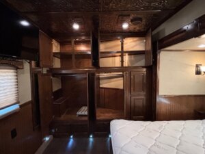 A wooden closet with multiple open cabinets and shelves is shown in a dimly lit room, resembling the cozy storage found in Living Quarters custom trailers. A white mattress sits nearby, with a window, closed blinds, and a TV mounted above. -Stillwater Trailer Sales