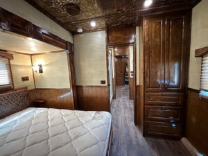 Interior of an RV or stock trailer showing a bedroom with an unmade mattress, wood-paneled walls and cabinets, small windows with blinds, and a view into a hallway with more cabinetry and detailed ceiling panels. Perfect for horse and livestock trailer sales. -Stillwater Trailer Sales