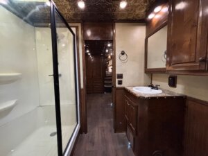A bathroom with a glass-enclosed shower on the left, a wooden vanity with a sink and granite countertop on the right, wood cabinets, and recessed ceiling lights—ideal for custom show trailers or stock trailers seeking upscale interiors. -Stillwater Trailer Sales