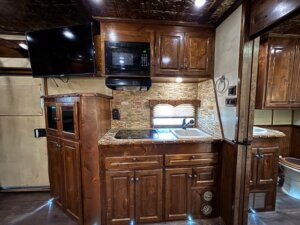 A compact RV kitchen with dark wood cabinets, a microwave, a small sink beneath a window, a wall-mounted TV, and part of a bathroom with toilet—all featured in our latest custom show trailers available at our trailer sales. -Stillwater Trailer Sales