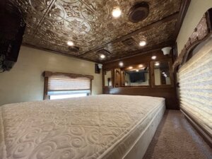 Interior view of a small bedroom with a patterned mattress, ornate tin ceiling, mirrors, wood trim, wall lights, and a window with a closed shade—much like the cozy spaces found in new and used trailers from top trailer sales. -Stillwater Trailer Sales