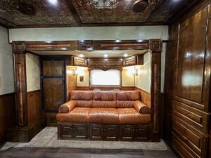 A luxurious RV interior, perfect for horse and livestock trailer sales enthusiasts, features a brown leather couch with rolled arms, wooden cabinetry, wall sconces, and a window with blinds set amid ornate woodwork and decorative trim. -Stillwater Trailer Sales