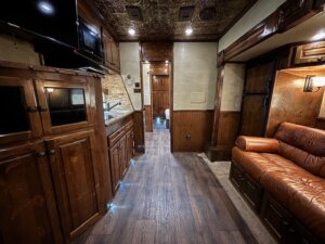 Interior of an RV or motorhome featuring dark wood cabinets, a small kitchen area, leather sofa, wood-style flooring, and recessed lighting. Perfect for Living Quarters custom designs often seen in horse and livestock trailer sales. TV is mounted above cabinets on the left. -Stillwater Trailer Sales