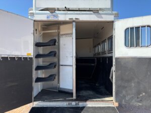 Open rear doors reveal saddle racks, bridle hooks, and a partitioned interior designed for horses. Custom Show Trailers available in trailer sales feature worn yet reliable floors and ample windows for comfort. -Stillwater Trailer Sales