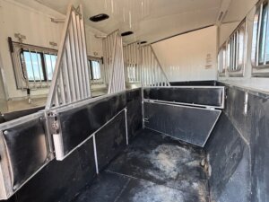 Interior of a custom show trailer featuring three divided stalls, padded walls, metal bars, and side windows for ventilation. The floor is covered in a dark, rubber-like material. Explore our trailer sales for more options. -Stillwater Trailer Sales