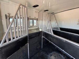 The image shows the interior of a horse trailer with three divided stalls, metal bars separating each section, and windows on the side. The walls and floor are lined with dark, durable material, offering quality similar to top Stock Trailers from trailer sales. -Stillwater Trailer Sales