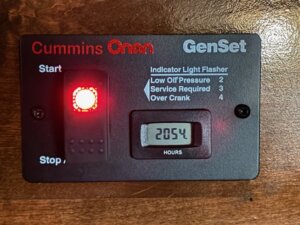 A Cummins Onan GenSet control panel in our Living Quarters custom Stock Trailers displays a red indicator light by Start and an hour meter reading 2054. Indicator instructions note low oil pressure, service required, and over crank conditions. -Stillwater Trailer Sales