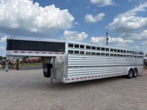 A large, silver livestock trailer with vented sides and multiple axles is parked on a gravel lot under a partly cloudy sky, showcasing quality options available through horse and livestock trailer sales. -Stillwater Trailer Sales