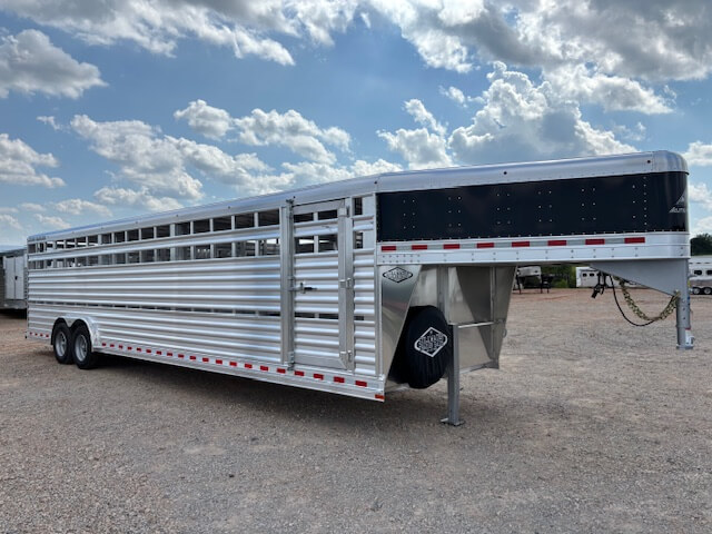 A large silver stock trailer with a black front section is parked on a gravel lot under a partly cloudy sky. Ideal for horse and livestock trailer sales, it features multiple vents, dual axles, and can be customized with Living Quarters. -Stillwater Trailer Sales