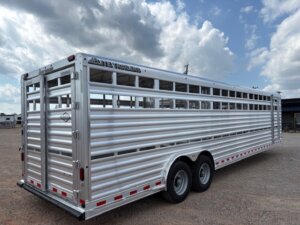 A large, silver stock trailer with two axles is parked on gravel under a partly cloudy sky. The trailer features slatted sides for ventilation and red and white reflectors—ideal for horse and livestock trailer sales. -Stillwater Trailer Sales