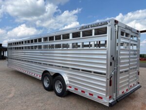 A large, silver livestock trailer with dual axles and ventilated sides is parked on gravel under a partly cloudy sky. Ideal for horse and livestock trailer sales, it features rear and side doors plus reflective red-and-white safety markings. -Stillwater Trailer Sales
