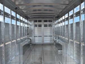 Interior view of an empty aluminum Stock Trailer with metal walls, floor, and benches on both sides. The ceiling and walls have ventilation holes; rear doors are closed. Find more new and used trailers at our trailer sales lot. Sky is visible through the vents. -Stillwater Trailer Sales