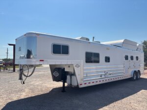 A large silver and white gooseneck horse and livestock trailer with multiple windows and vents is parked on a gravel lot under a clear sky, showcasing quality options available at our horse and livestock trailer sales. -Stillwater Trailer Sales