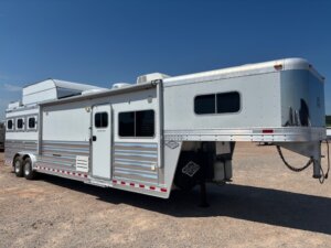 A large silver gooseneck horse trailer with Living Quarters custom design is parked on gravel under a clear blue sky. The trailer features multiple windows, roof vents, and spacious storage compartments—a great example of modern Trailer sales. -Stillwater Trailer Sales