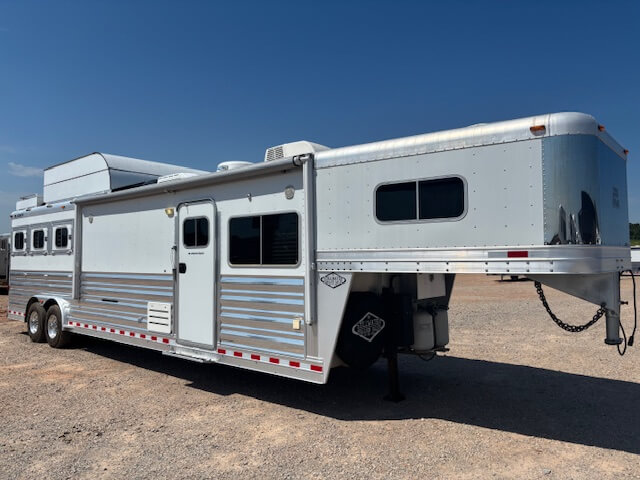 A large silver gooseneck horse trailer with living quarters is parked on gravel under a clear blue sky. Perfect for buyers seeking new and used trailers, this trailer features multiple windows, vents, and air conditioning units on the roof. -Stillwater Trailer Sales