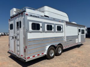 A large silver and white Platinum horse trailer is parked on gravel under a clear blue sky. Featuring multiple windows, two axles, a rear door with ladder, it’s ideal for those seeking new and used trailers or custom living quarters options. -Stillwater Trailer Sales