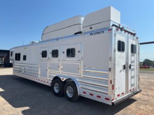 A large, silver gooseneck horse trailer with multiple doors and windows is parked on gravel. Perfect for stock trailers or horse and livestock trailer sales, it features dual axles, ventilation units, and “Platinum” branding under a clear blue sky. -Stillwater Trailer Sales