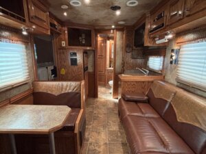 The image shows the interior of an RV with brown wood cabinets, a booth-style dining area, leather sofa, tile flooring, and a view of the kitchenette and bathroom—perfect inspiration for trailer sales or those seeking custom show trailers. -Stillwater Trailer Sales