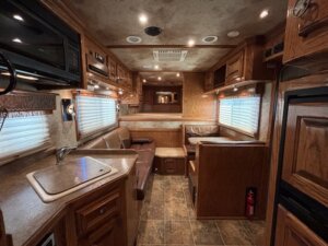 The interior of an RV featuring a compact kitchen with a sink and microwave, a leather sofa, dining booth, wood cabinetry, tiled flooring, and a small fire extinguisher by the refrigerator—perfect for those exploring new and used trailers. -Stillwater Trailer Sales