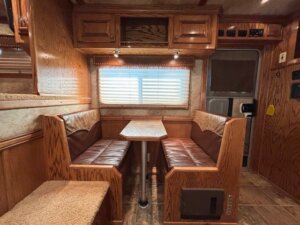 A wooden RV dinette with two brown leather benches facing each other, a rectangular table in between, cabinets above, and a window with closed blinds and two overhead lights—perfect for relaxing during your new and used trailer adventures. The RV door is visible to the right. -Stillwater Trailer Sales