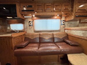 A brown leather couch with two matching armrest cushions sits against a wood-paneled wall under a window with closed blinds in a cozy RV interior—perfect for relaxing during horse and livestock trailer sales or while browsing new and used trailers. Cabinets and a sink are nearby. -Stillwater Trailer Sales