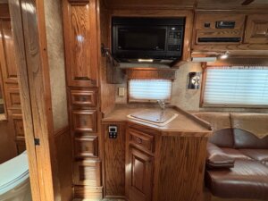 A compact RV kitchen with wood cabinetry, a built-in microwave above a small sink, and a window with a blind. Perfect for Living Quarters custom setups or Custom Show Trailers for horse and livestock trailer sales. Corner sofa and steps add to its charm. -Stillwater Trailer Sales
