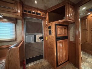 Interior view of an RV with wood-paneled cabinets and trim, a small dining area to the left, metal door with window covering in the center, and built-in refrigerator on the right—perfect for those interested in new and used trailers or exploring trailer sales. -Stillwater Trailer Sales