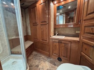 RV bathroom with wood cabinets, a glass-enclosed shower on the left, small bench, sink with mirror above, countertop, and tiled flooring. A portion of a toilet is visible—perfect for those exploring new and used trailers at your local trailer sales. -Stillwater Trailer Sales