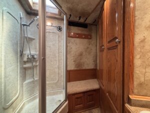 A small bathroom features a glass-enclosed shower with built-in shelves and a handheld showerhead, a cushioned bench, wooden cabinets, and coat hooks on the wall under a skylight—ideal touches for those visiting after horse and livestock trailer sales. -Stillwater Trailer Sales