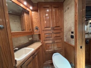 A small bathroom with wood cabinetry, a white sink, a built-in mirror, and beige textured walls—an ideal feature in custom show trailers designed for horse and livestock trailer sales. Recessed lighting is above the mirror; electrical switches are on the wall. -Stillwater Trailer Sales