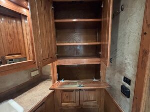 A wooden cabinet with open doors and shelves is built into a corner above a countertop, resembling the organized spaces found in living quarters custom horse and livestock trailer sales. Cabinets below, outlets, and switches are near the right; a mirror and sink on the left. -Stillwater Trailer Sales
