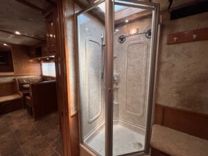 A glass-enclosed shower with a beige interior stands in the corner of an RV bathroom. The RV’s living area, featuring a dining booth and wood cabinets, showcases Living Quarters custom options popular in trailer sales. -Stillwater Trailer Sales