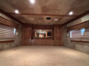 The interior of this RV bedroom from our Trailer sales lineup features carpet flooring, wooden cabinets, a large mirror, and windows with striped blinds—perfect for those seeking Living Quarters custom comfort on the road. Ceiling lights are turned on. -Stillwater Trailer Sales