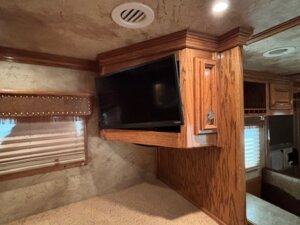 A flat-screen TV is mounted at an angle on a wooden cabinet in an RV interior. The area, perfect for relaxing after checking out horse and livestock trailer sales, features beige walls, a window with a blind and valance, and recessed ceiling lights. -Stillwater Trailer Sales