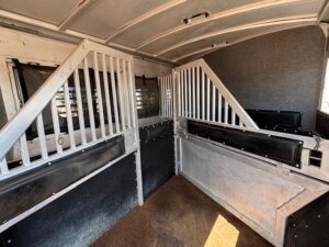 Interior of a horse trailer with divided stalls, metal bars, padded walls, and windows allowing light to enter. The textured brown floor reflects the quality seen in premium Trailer sales and Custom Show Trailers. -Stillwater Trailer Sales