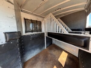 Interior of a horse and livestock trailer sales model with padded walls, a window with protective grill, grated divider, and brown flooring—designed for transporting animals safely. -Stillwater Trailer Sales