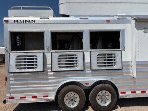 A silver horse trailer with three open windows, each featuring metal bars and ventilation slats. Ideal for horse and livestock trailer sales, it has two axles, four wheels, and the PLATINUM brand name displayed at the top. -Stillwater Trailer Sales