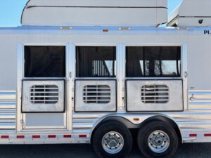 Side view of a silver horse trailer with two large open windows featuring metal bars and mesh coverings, dual wheels underneath—perfect for those seeking new and used trailers or expert trailer sales. Part of another trailer is visible at the top right. -Stillwater Trailer Sales