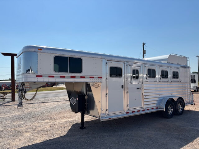 A silver Elite 4 Horse gooseneck trailer with four windows and two axles is parked on gravel under a clear blue sky. -Stillwater Trailer Sales