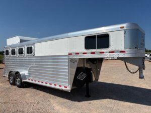 An Elite 4 Horse silver gooseneck livestock trailer with four windows and two axles is parked on a gravel lot under a clear blue sky. -Stillwater Trailer Sales