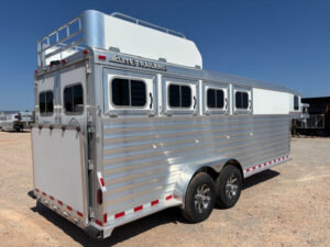 A silver and white Elite 4 multi-horse trailer with four windows on the side, dual axles with chrome wheels, and a roof rack, parked on gravel under a clear blue sky. Perfect for transporting your Horse Mustang in style. -Stillwater Trailer Sales