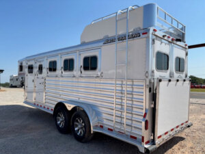 A silver Elite 4 Horse trailer with four side windows, two rear doors, dual axles, and a roof ladder is parked on gravel under a clear sky. The Mustang-inspired trailer features “Elite Trailers” branding on top. -Stillwater Trailer Sales